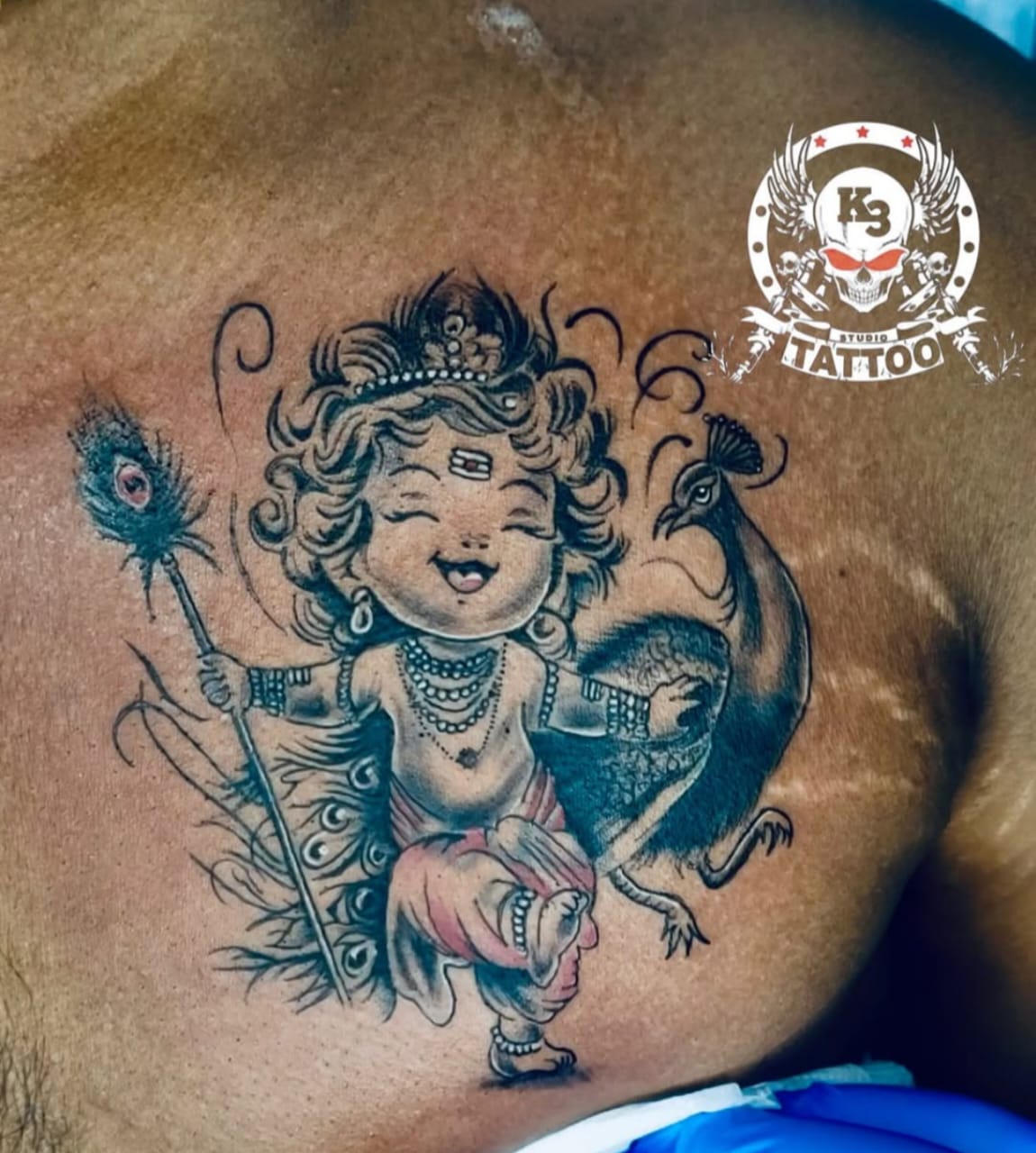 Colorful illustrative tattoo of baby Krishna with a peacock on the chest